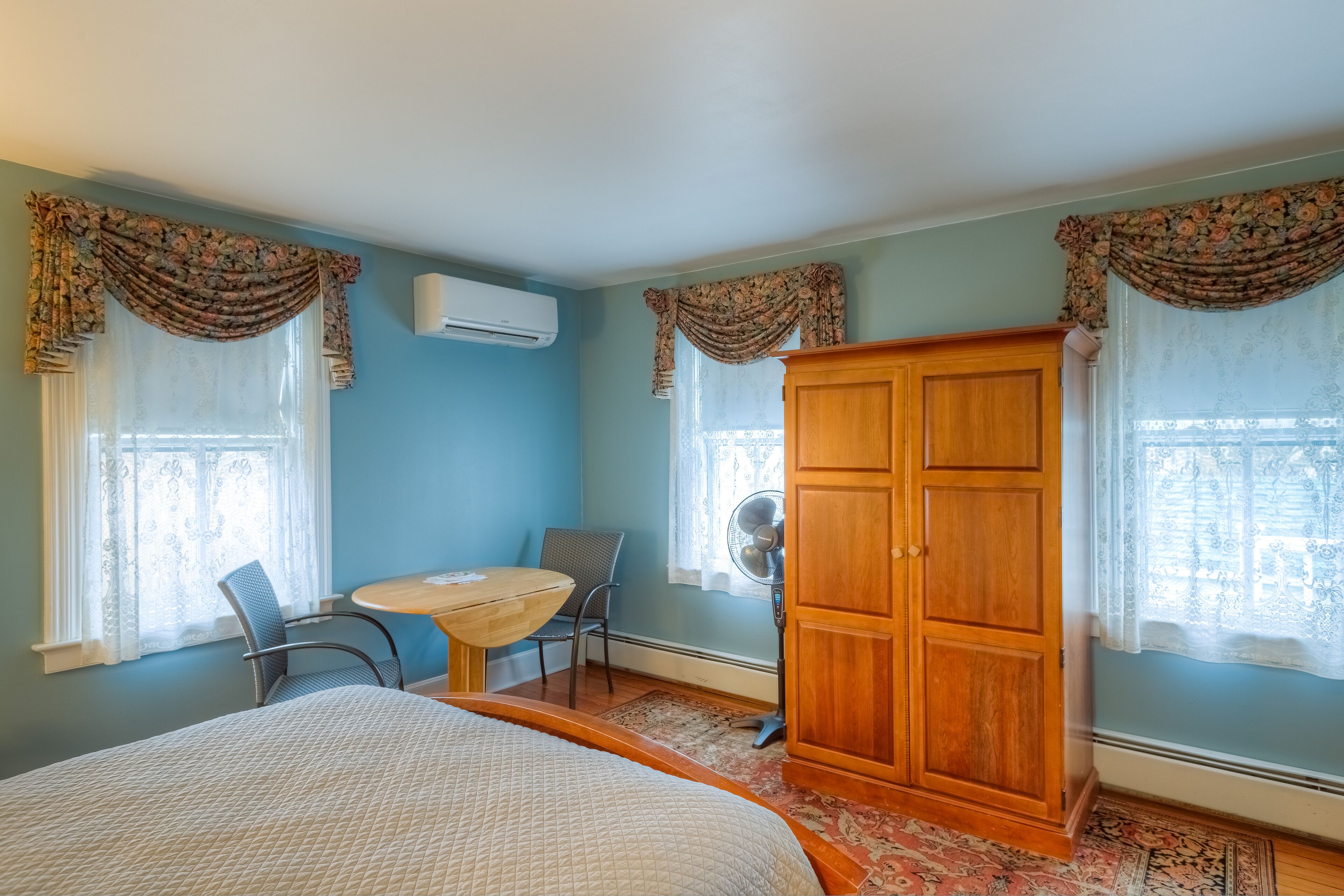 A bedroom with a bed, a wooden wardrobe, a desk with a chair, and two windows with curtains.