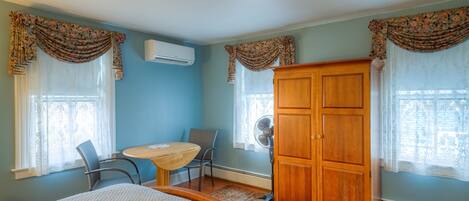 A bedroom with a bed, a wooden wardrobe, a desk with a chair, and two windows with curtains.