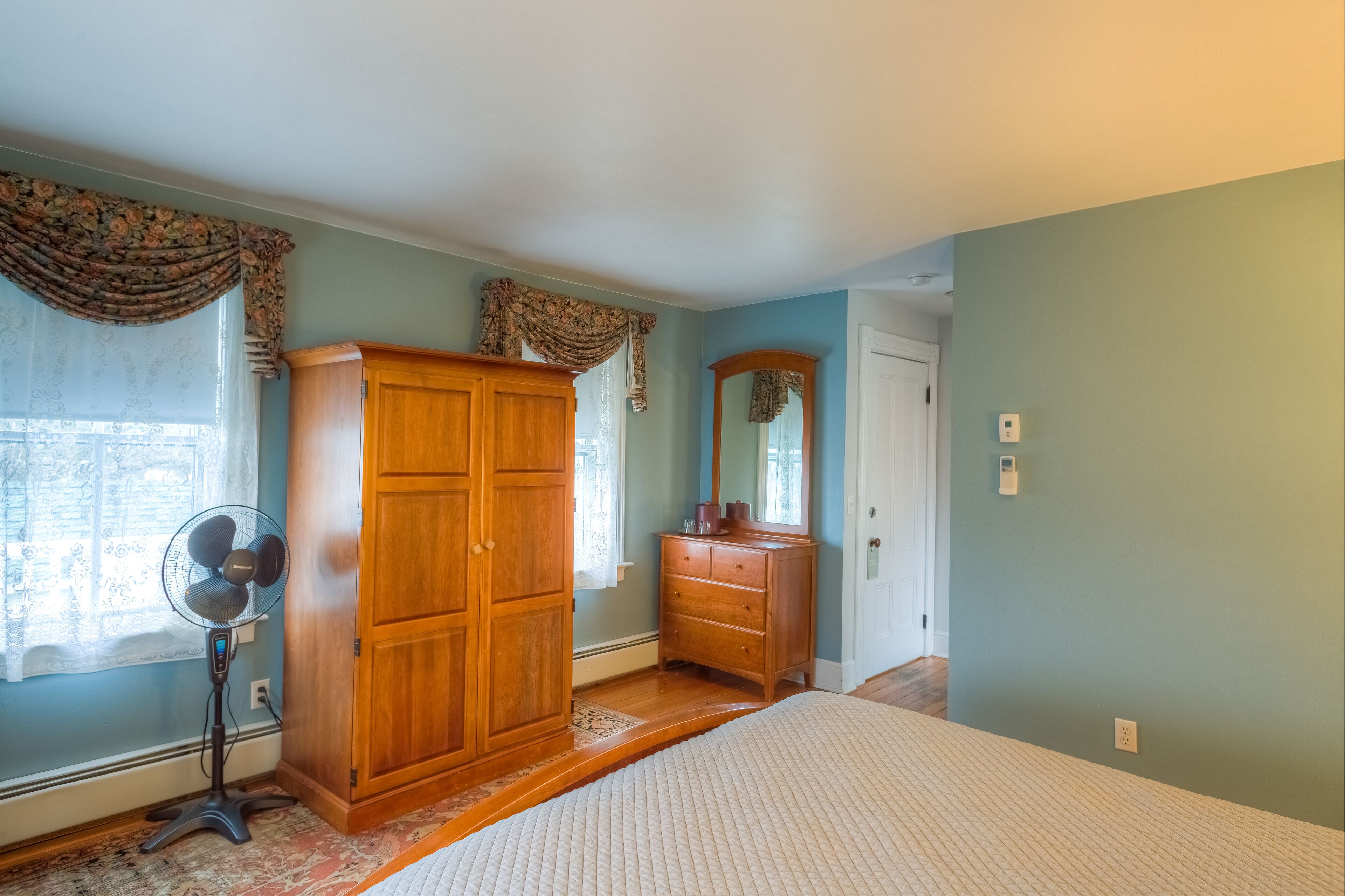 Une chambre avec une armoire en bois, une commode, un miroir et un ventilateur sur pied.