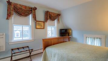 A bedroom with a bed, a bench, a TV, and two windows with curtains.