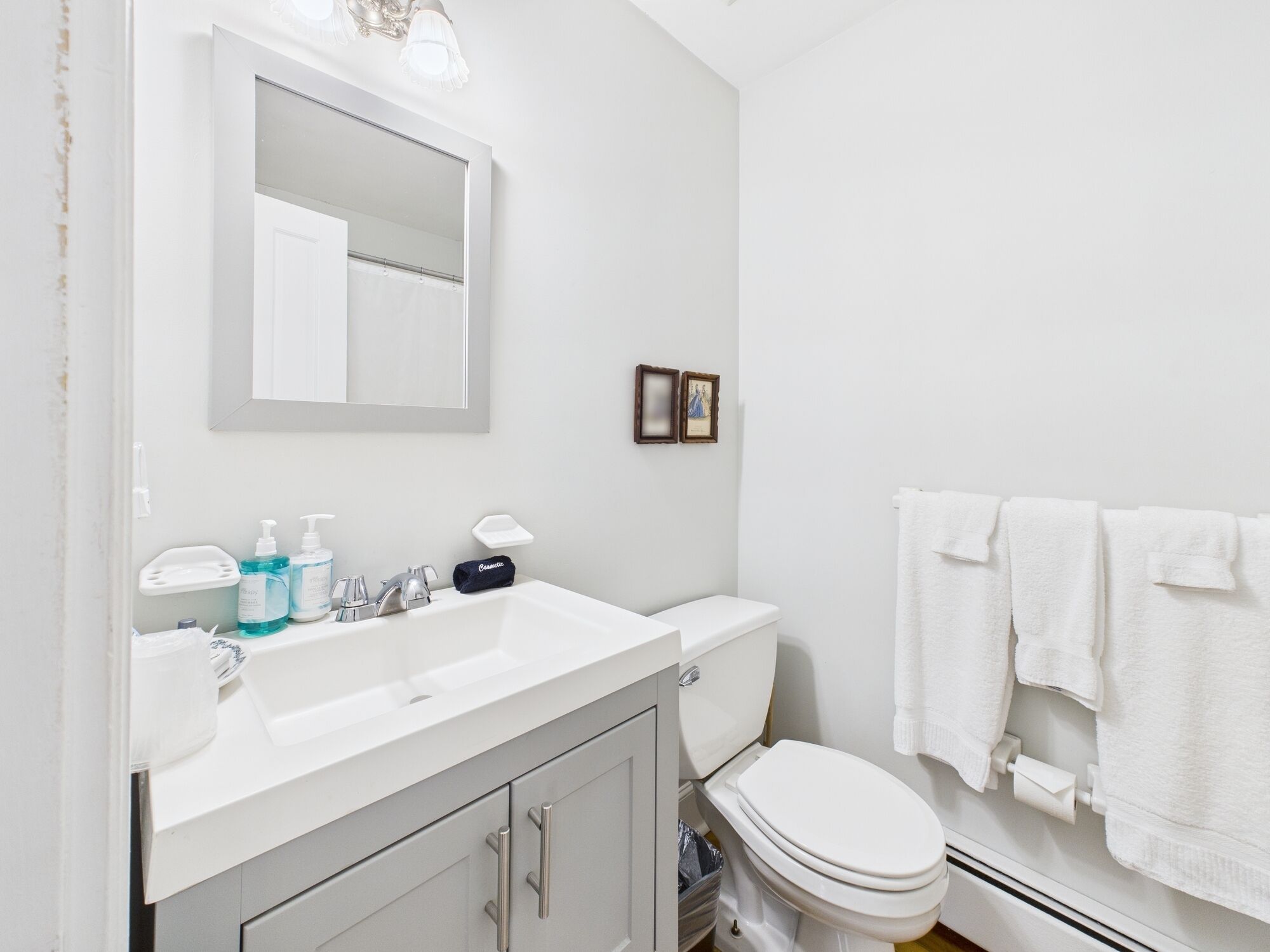 A bathroom with a white sink, mirror, and toilet. There are toiletries on the sink, a towel rack with towels, and a small picture frame on the wall.