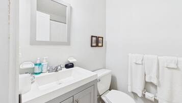 A bathroom with a white sink, mirror, and toilet. There are toiletries on the sink, a towel rack with towels, and a small picture frame on the wall.