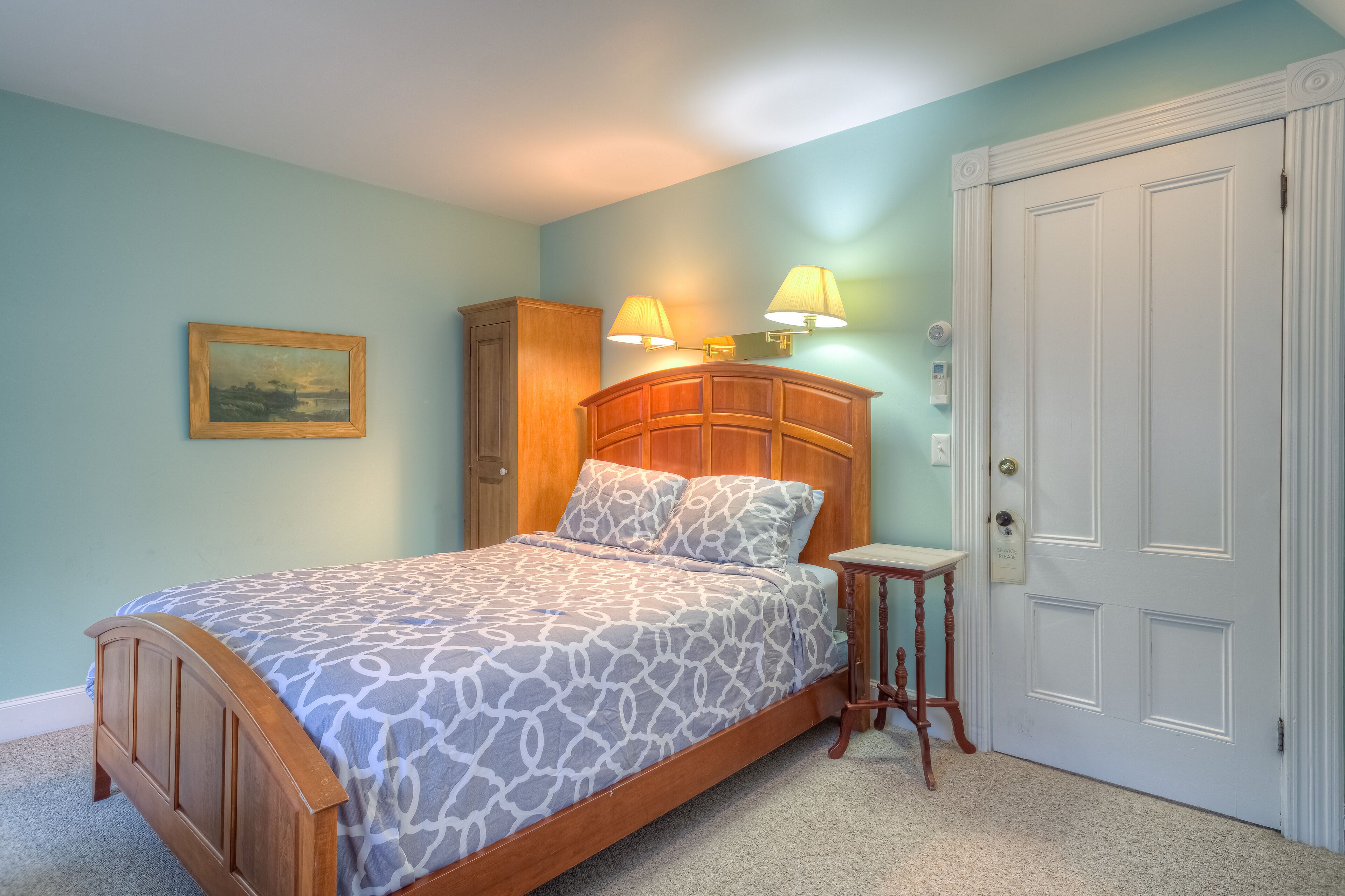 A bedroom with a wooden bed, two bedside lamps, a nightstand, a door, and a painting on the wall.
