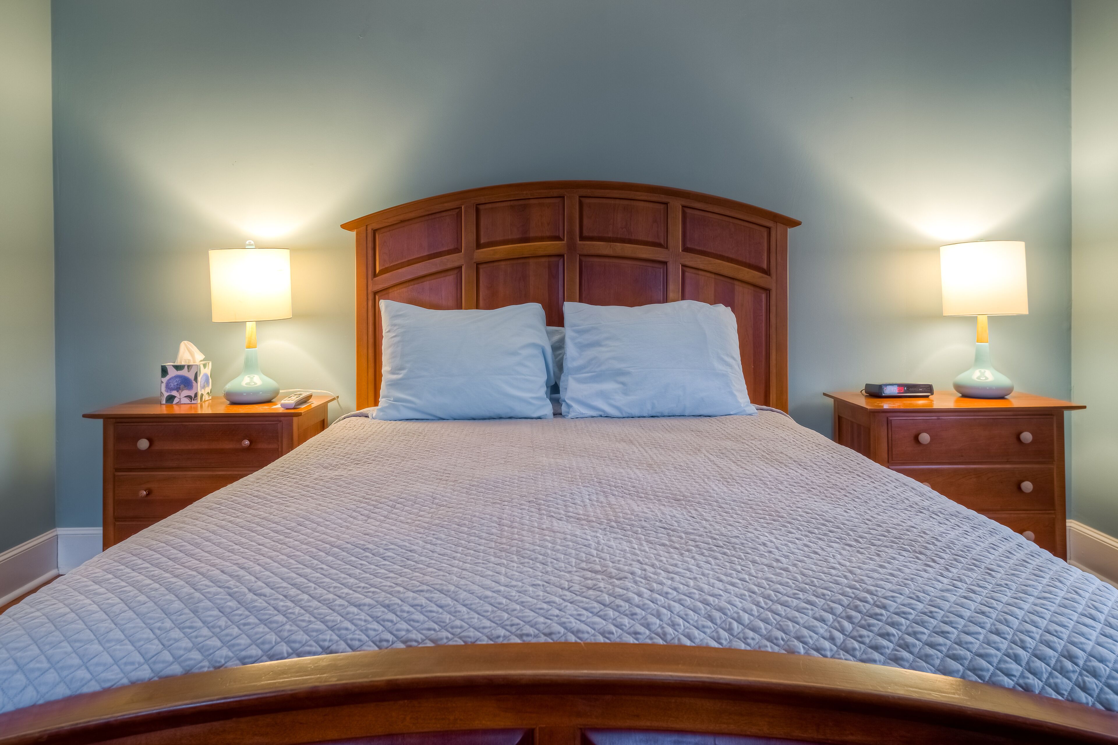 Una cama bien tendida con cabecera de madera, dos mesitas de noche con lámparas y una mesita con una caja de pañuelos y un teléfono.