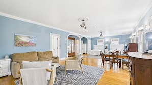 A spacious living room with a sofa, a dining table with chairs, a kitchen area, and a hallway leading to bedrooms. - Dodge Cottage (Block Island)