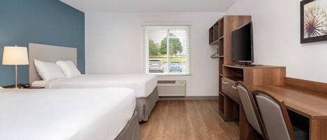 A hotel room with a bed, bedside table, lamp, TV, desk, chair, and a window with blinds.