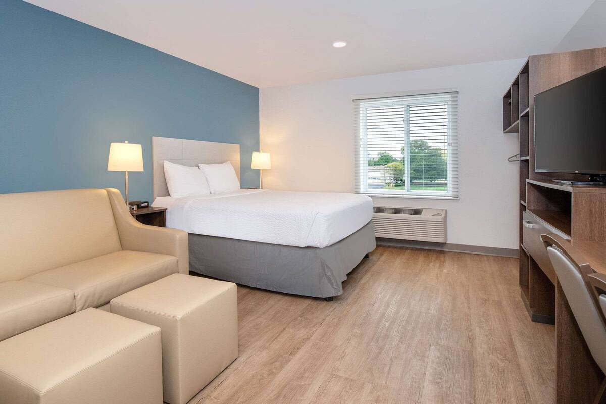 A hotel room with a bed, a sofa, a TV, and a window with blinds.