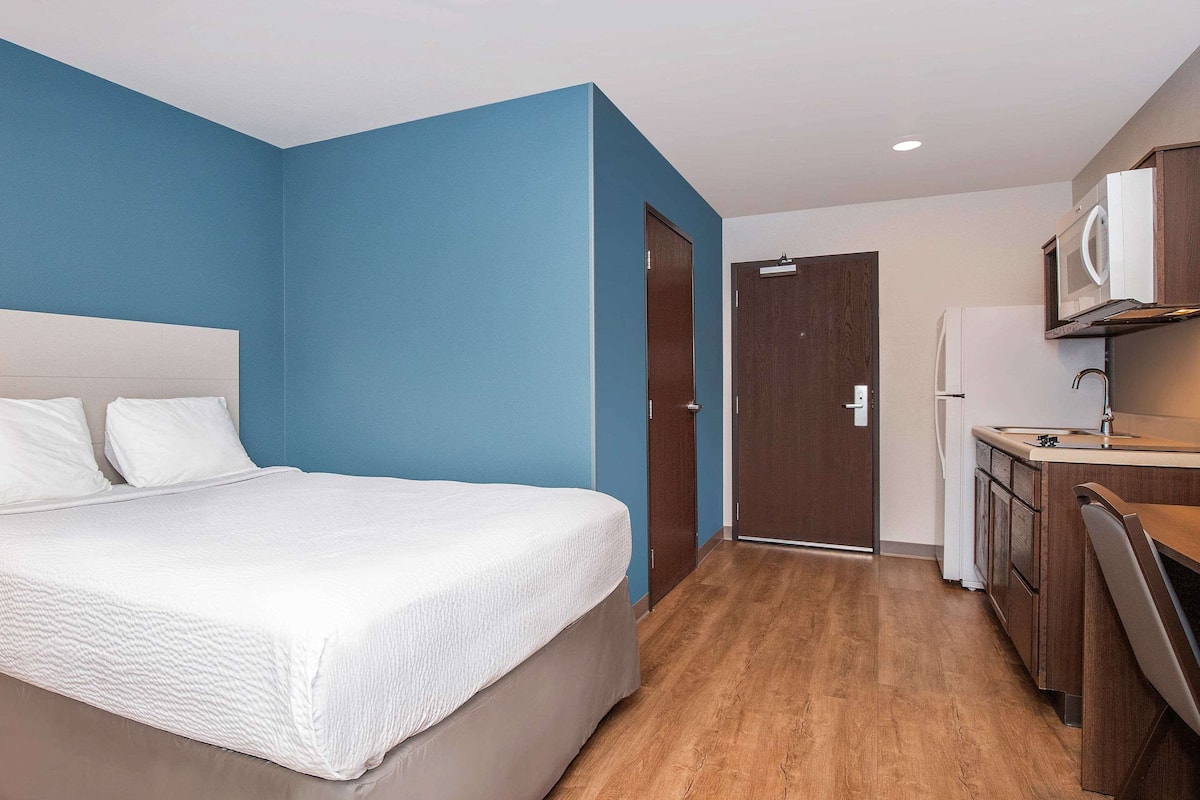 A hotel room with a bed, a kitchenette, and a door leading to another room.