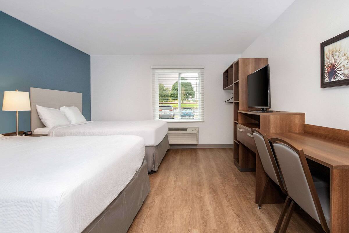 A hotel room with two beds, a desk with chairs, a TV, and a window with blinds.