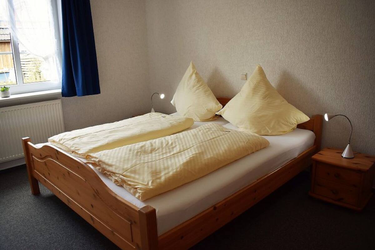 A wooden bed with a white mattress and two pillows, a bedside table with a lamp, and a window with blue curtains.