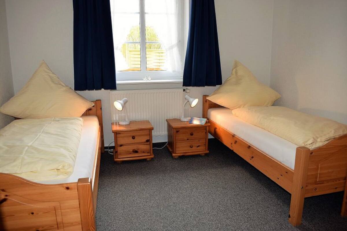 A small, cozy room with two wooden beds, each with a pillow and a bedside table with a lamp.