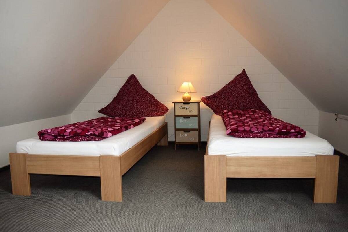 A small, triangular attic room with two wooden beds, each with a red patterned blanket and pillow. There is a nightstand with a lamp between the beds.