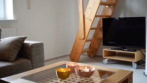 A living room with a sofa, a glass-top coffee table with candles, a wooden ladder, and a TV on a stand. - Haus Uthlande (Dagebuell)