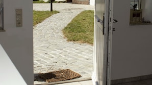 Una puerta abierta que da a un patio con un camino empedrado, una puerta de madera y un pequeño ventanal con un arreglo floral.