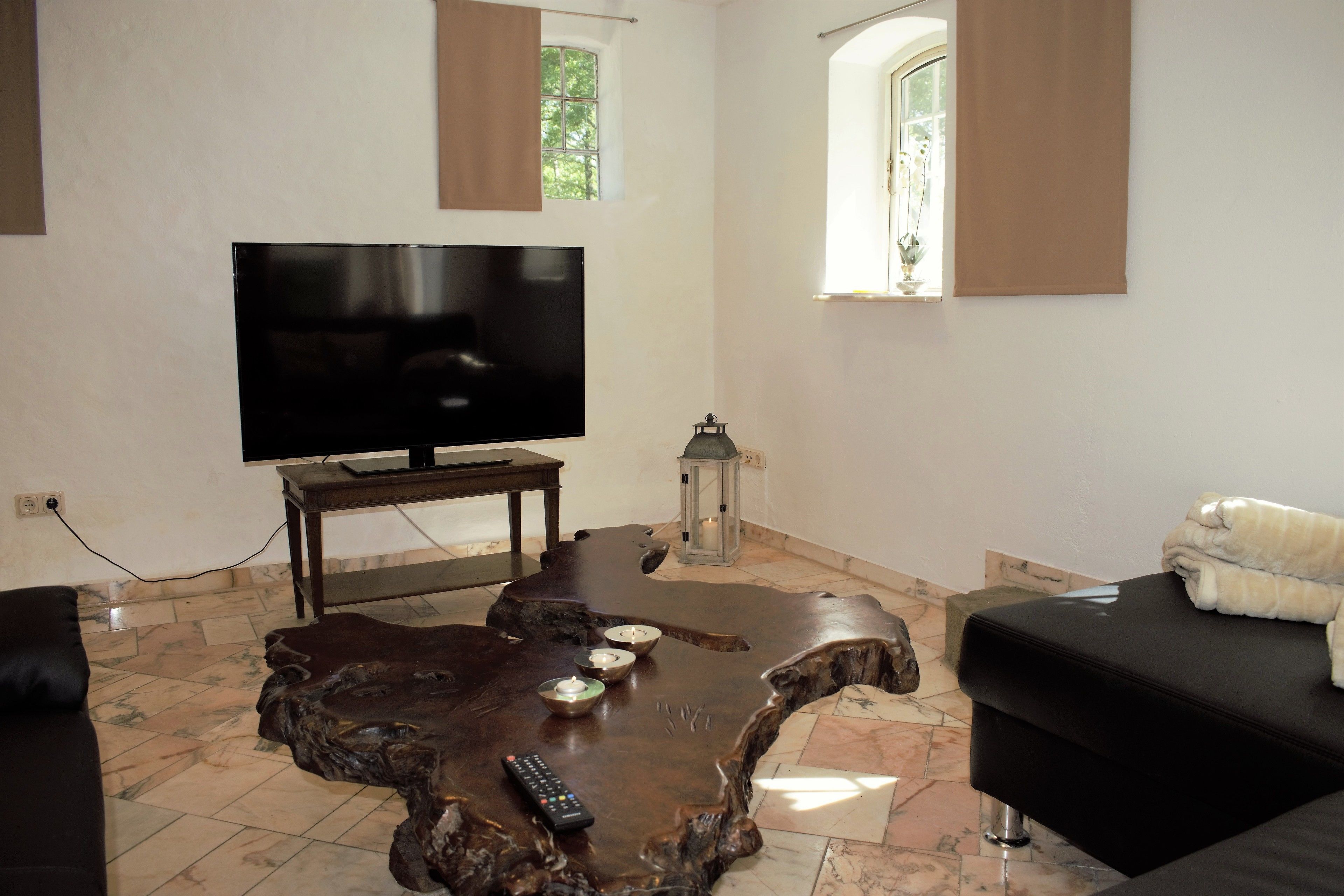 Una sala de estar con un televisor de pantalla plana grande, una mesa de centro de madera con velas, un sofá negro y una ventana con cortinas.