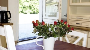 Una mesa de comedor de madera con un jarrón blanco de rosas rojas, sillas blancas, una cafetera y un armario con puertas de vidrio.
