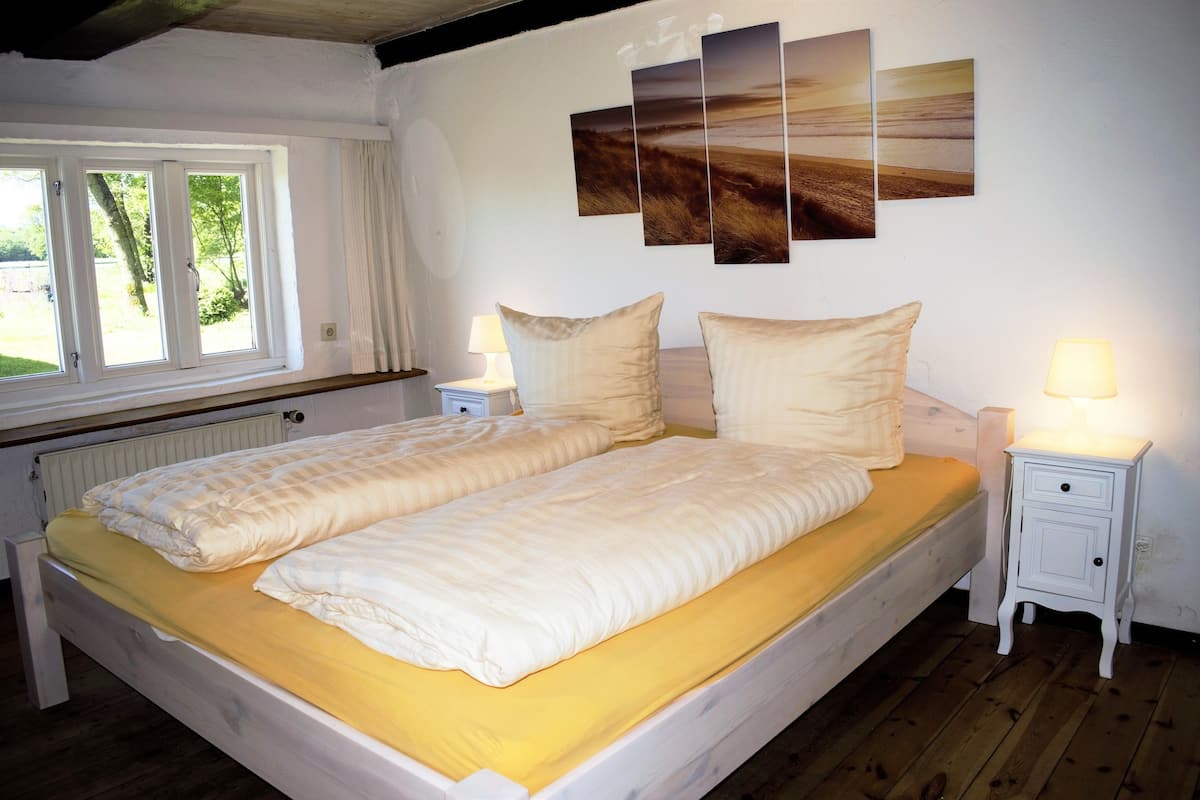 Una cama doble con una colcha amarilla, dos mesitas de noche con lámparas, un ventanal con cortinas y una pared con tres cuadros de paisajes.