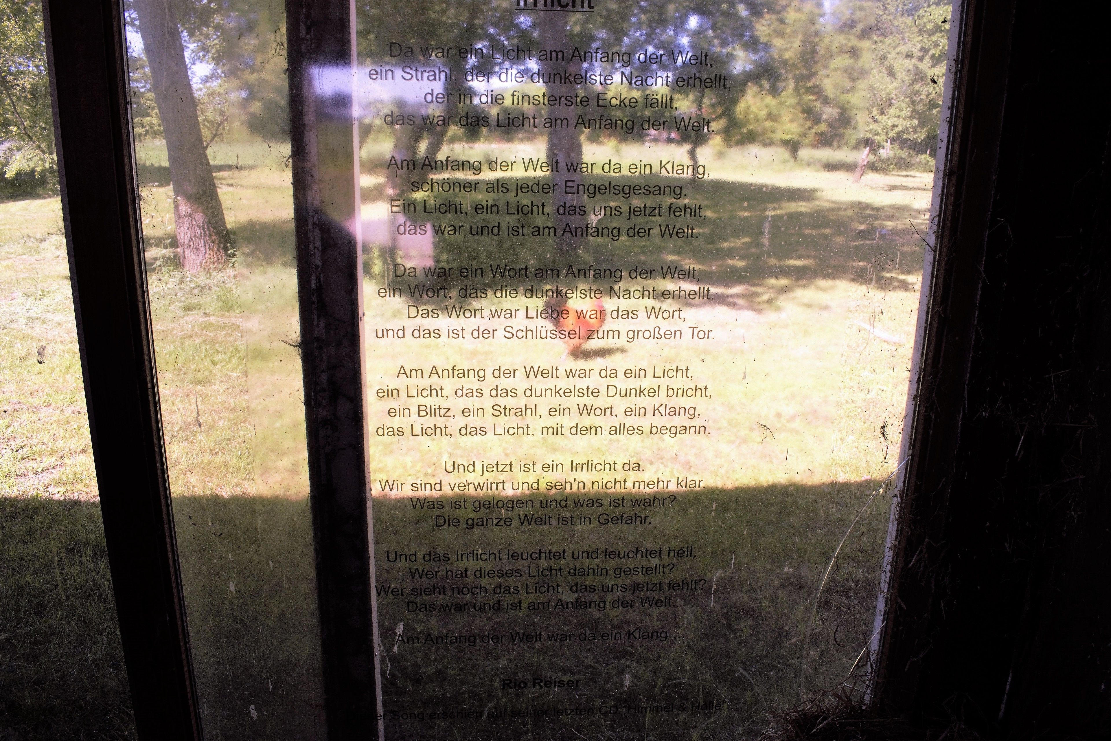 Ein Fenster mit Blick auf eine Wiese und Bäume, durch das ein Gedicht zu sehen ist.