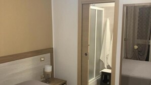 Habitación de hotel con dos camas, una mesita de noche con lámpara, un espejo y zona de ducha.