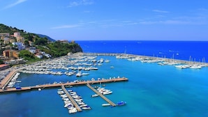 Un puerto deportivo con numerosas embarcaciones amarradas, un muelle que se adentra en el agua y edificios en una colina al fondo.