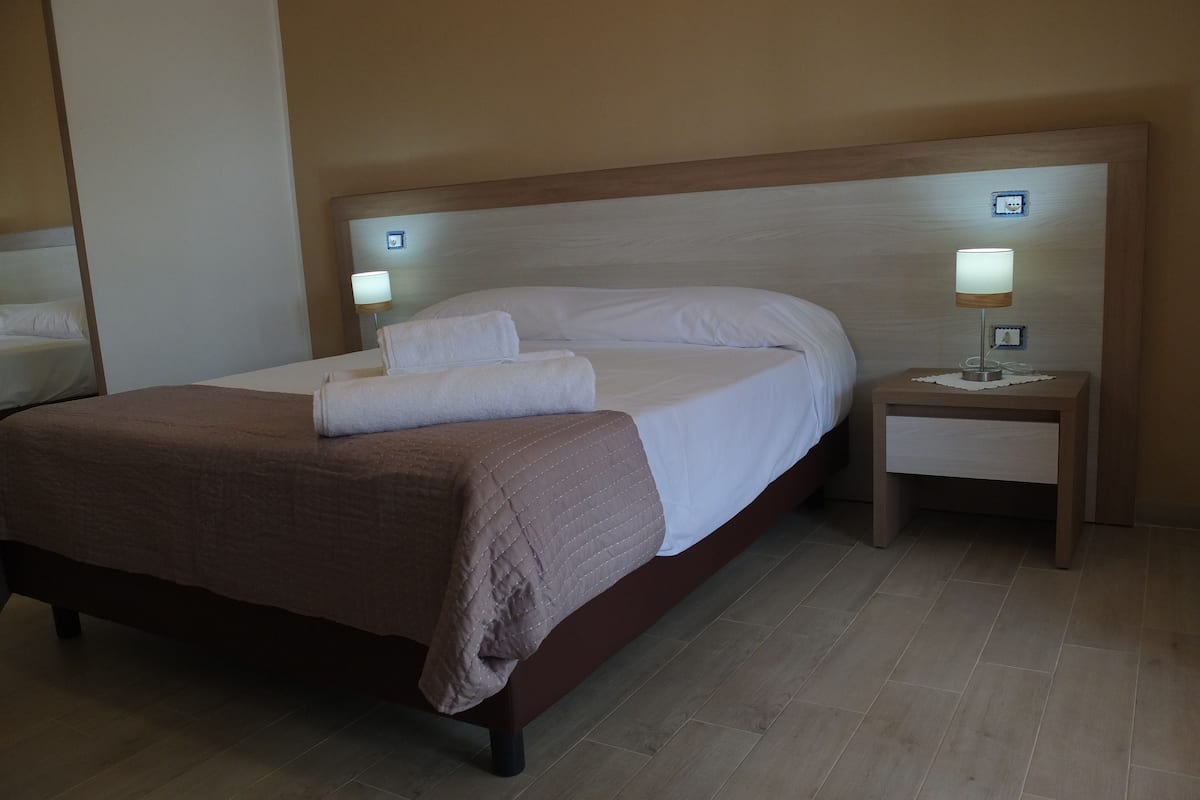 A neatly made bed with a brown bedspread and white linens, a bedside table with a lamp, and a wall-mounted light fixture.