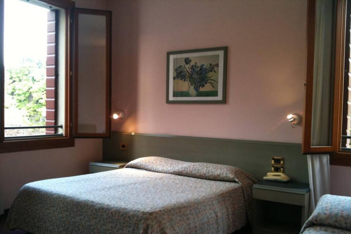 A bedroom with two beds, a nightstand, a bedside table, a framed floral artwork, and a window with blinds.