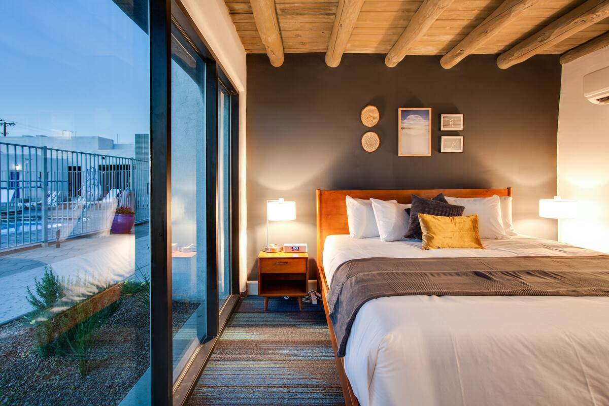 Ein Hotelzimmer mit einem großen Bett, einem Nachttisch, einem Fenster mit Blick auf die Stadt und einer Holzbalkendecke.