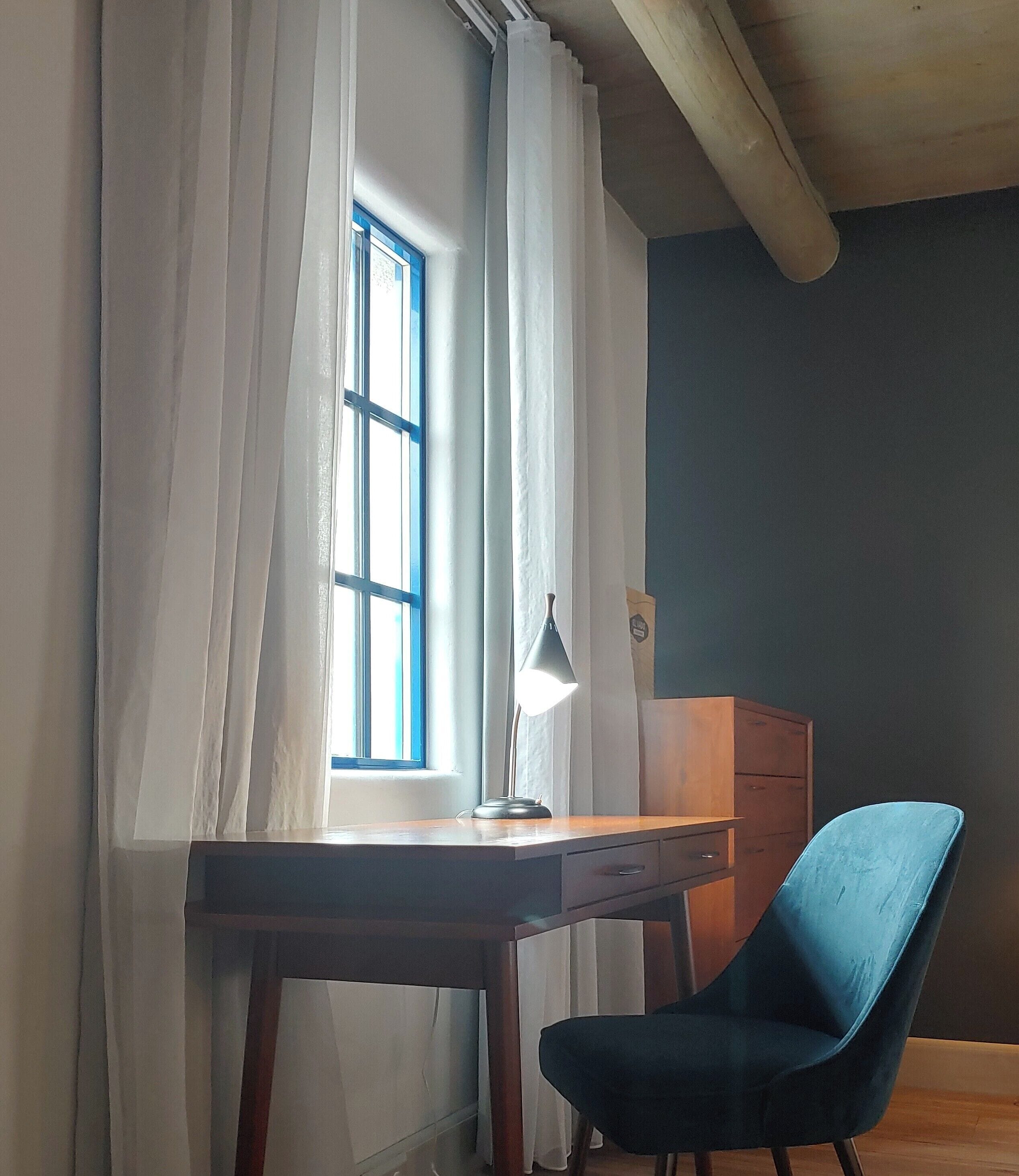 A room with a wooden desk, a blue chair, a lamp, and a window with white curtains.