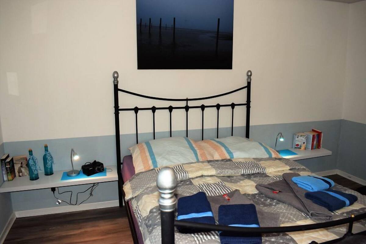 A neatly made bed with a metal headboard, a bedside table with a lamp, and a shelf with books and a small plant.
