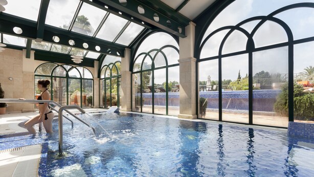 An indoor swimming pool with clear glass walls, a tiled floor, and a person in a swimsuit. - La Sella Premium Lifestyle Apartments (Denia)