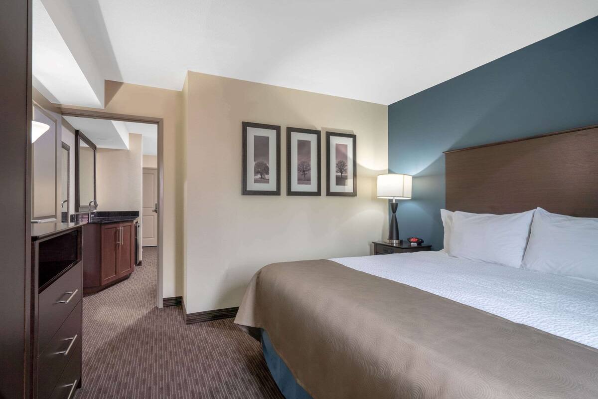 A hotel room with a large bed, a nightstand with a lamp, a dresser, and a mirror. There are three framed pictures on the wall.