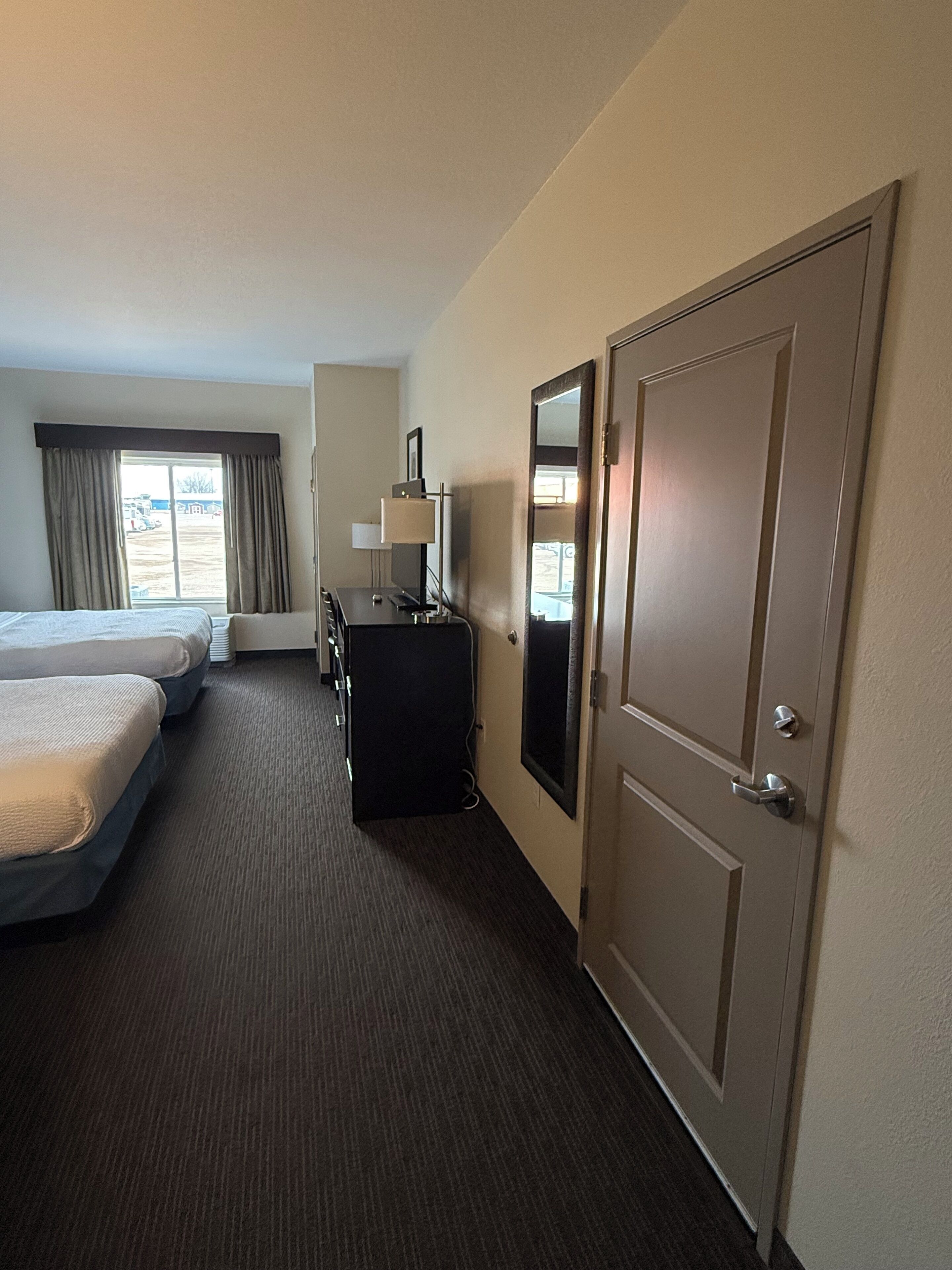 A hotel room with two beds, a desk, a chair, a lamp, and a door.