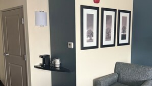 Habitación de hotel con un sofá gris, una pequeña mesa con cafetera, un cesto de basura y una puerta. Hay tres cuadros en la pared.