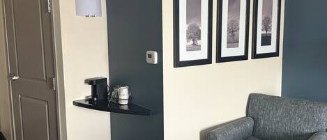 A hotel room with a grey sofa, a small table with a coffee maker, a trash bin, and a door. There are three framed pictures on the wall.