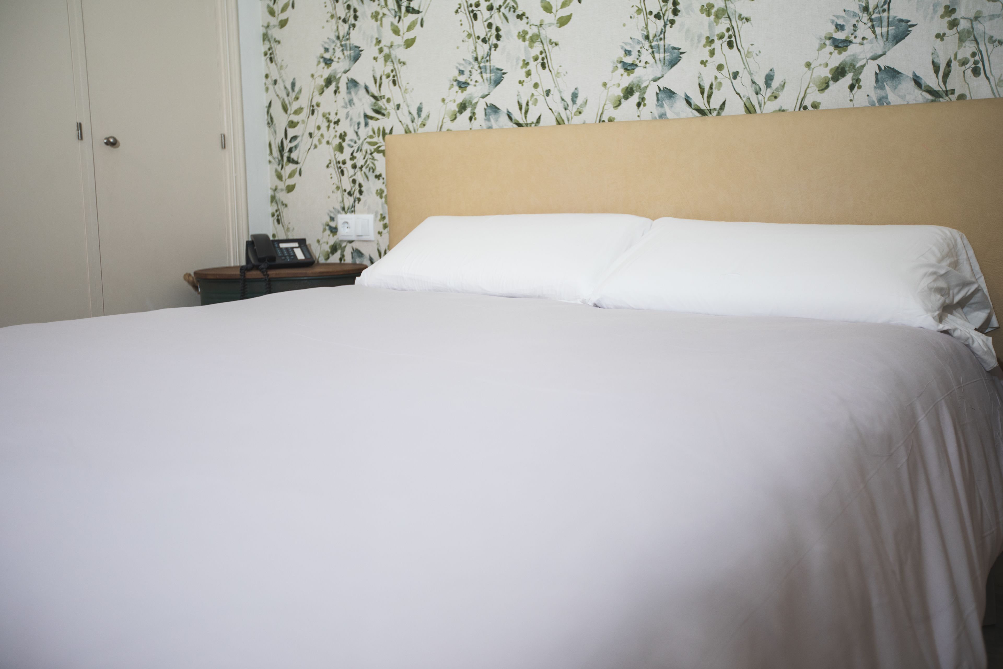 A neatly made bed with white linens in a room with floral wallpaper and a beige headboard.