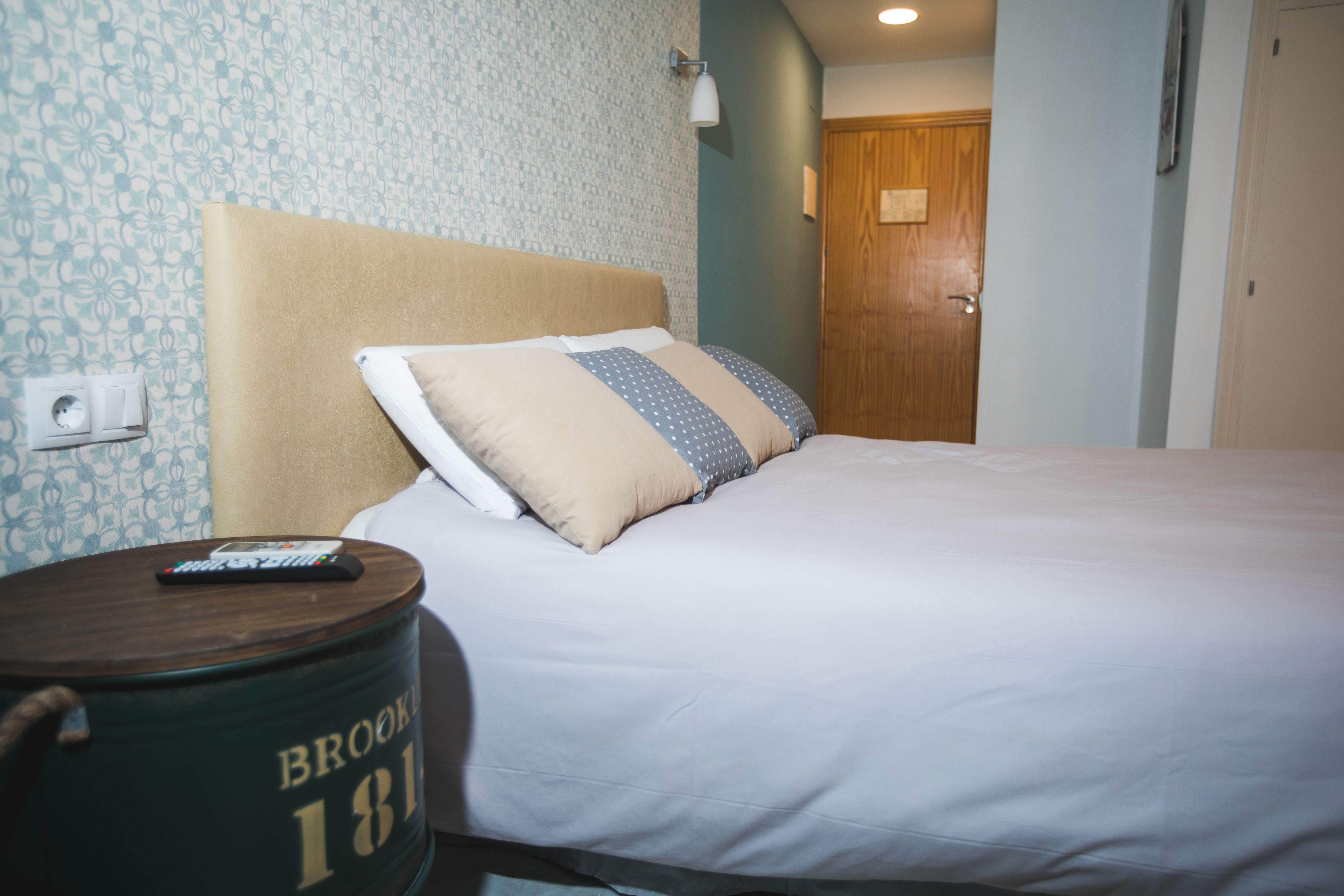 A neatly made bed with a wooden headboard, a bedside table with a remote control, and a wall with a patterned wallpaper.