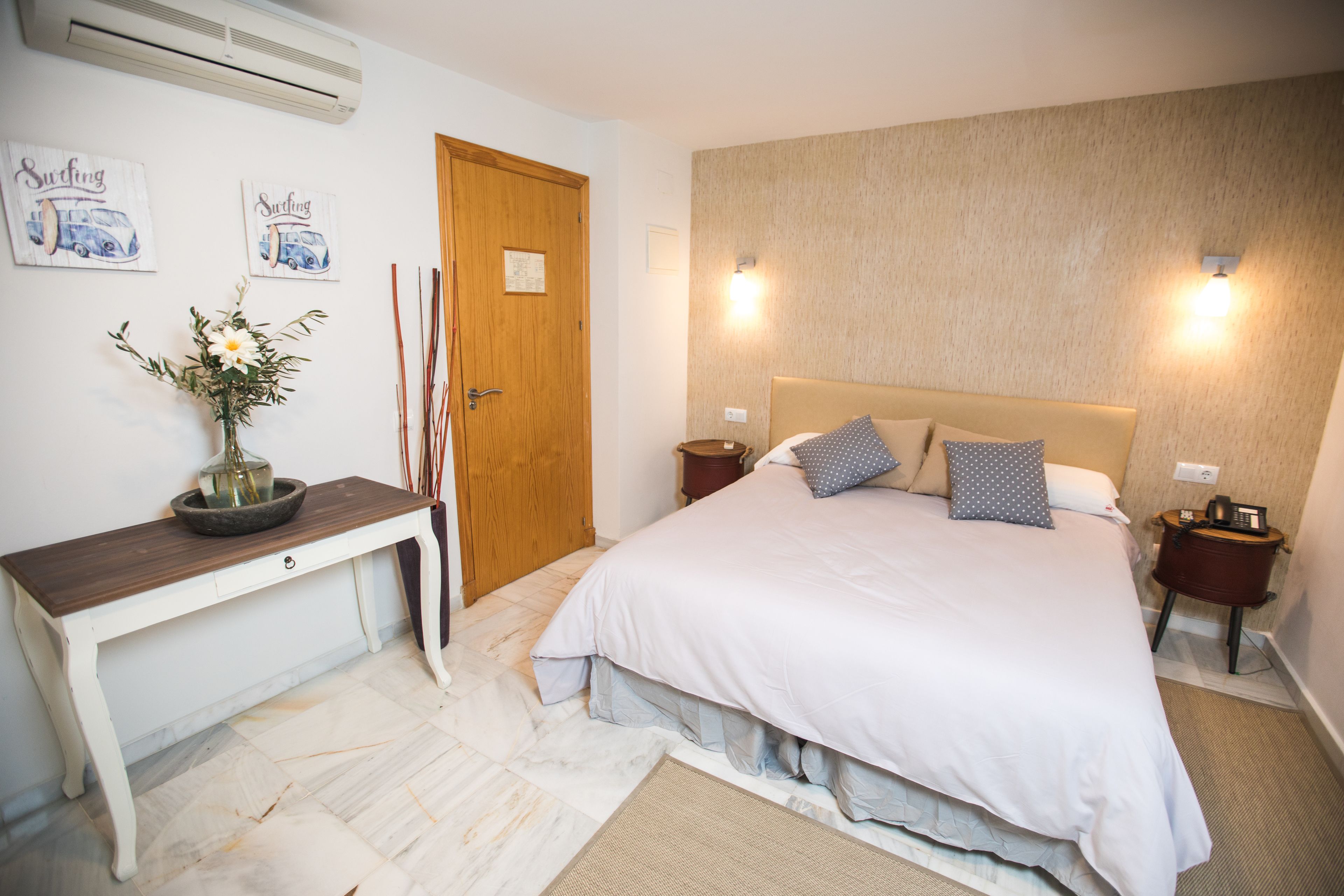 Un dormitorio con cama, mesitas de noche, un escritorio de madera, un jarrón con flores y decoración en la pared.