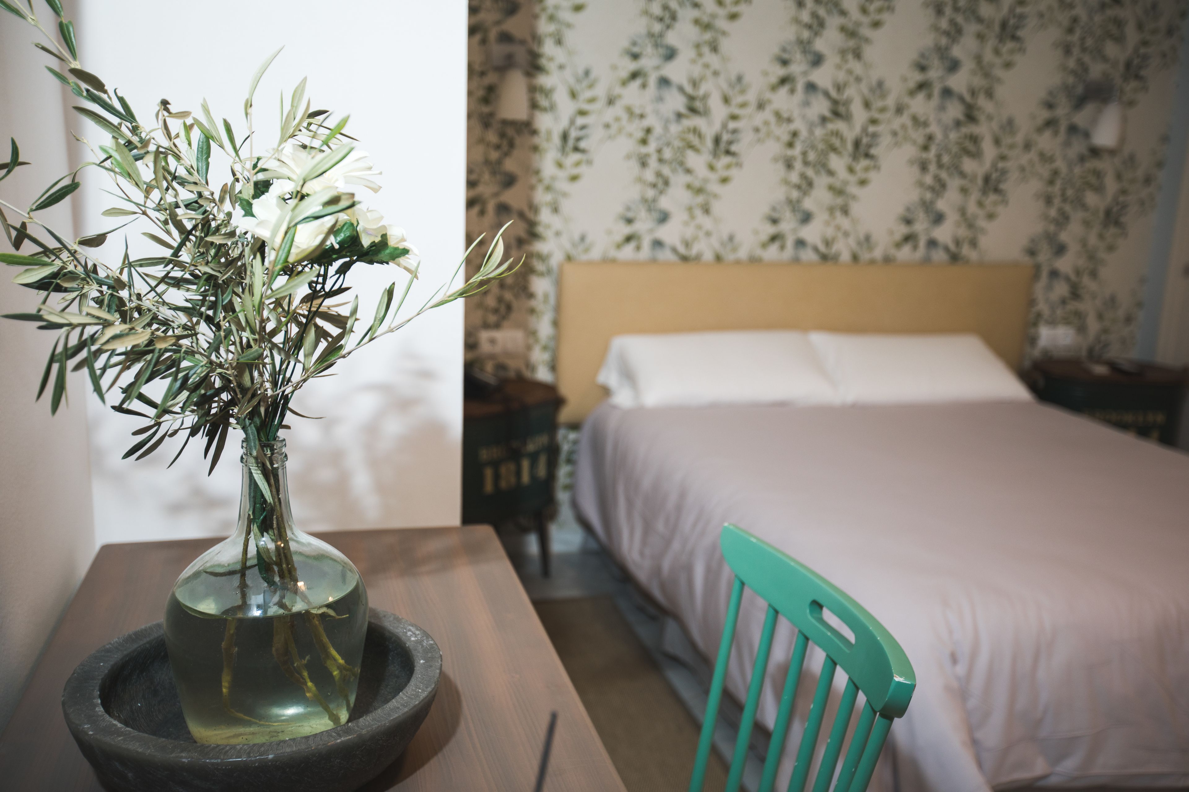 A room with a bed, a nightstand, a green chair, and a vase with flowers on a table.