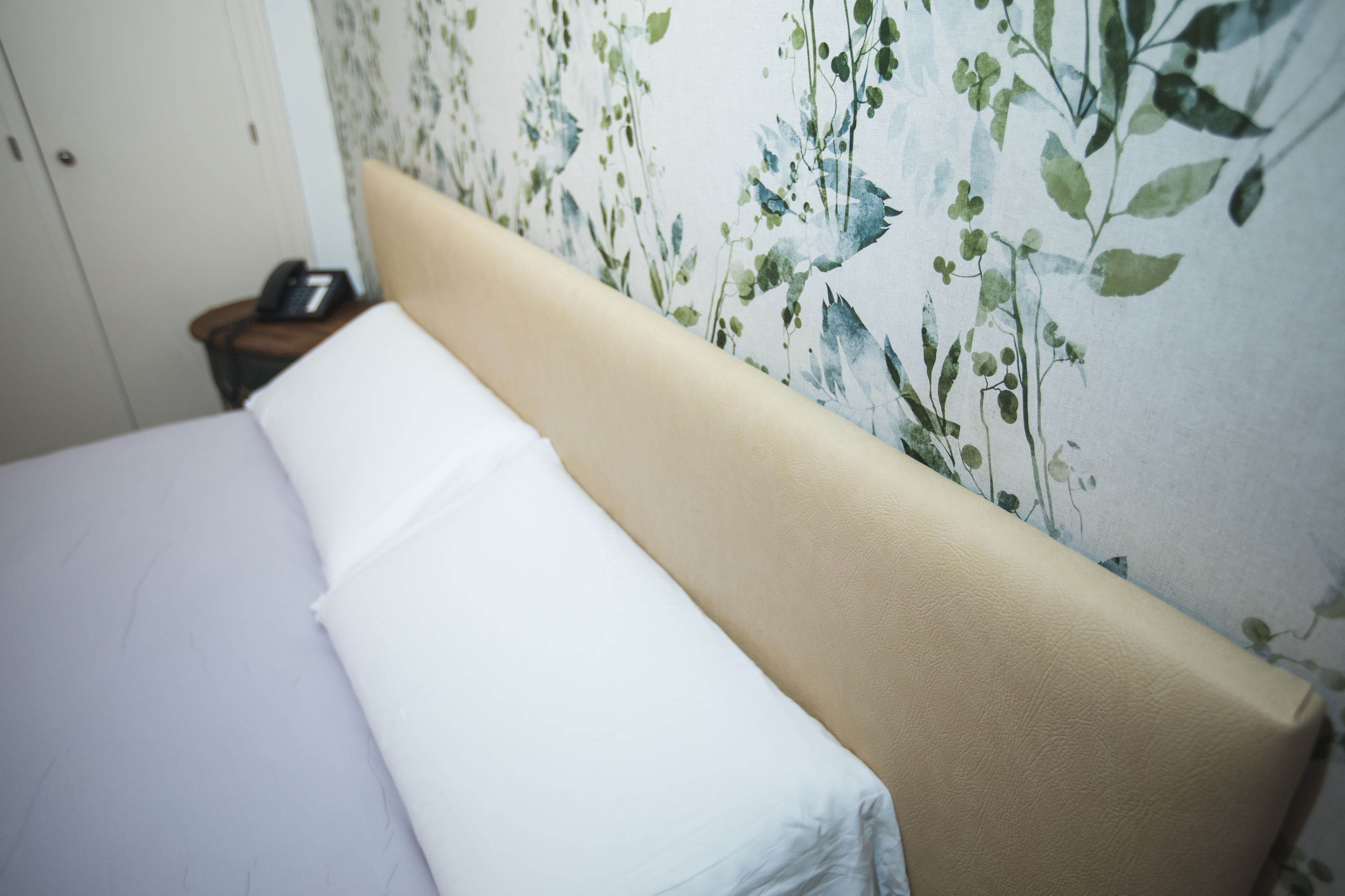A bed with white linens and a beige headboard against a floral wallpaper backdrop.