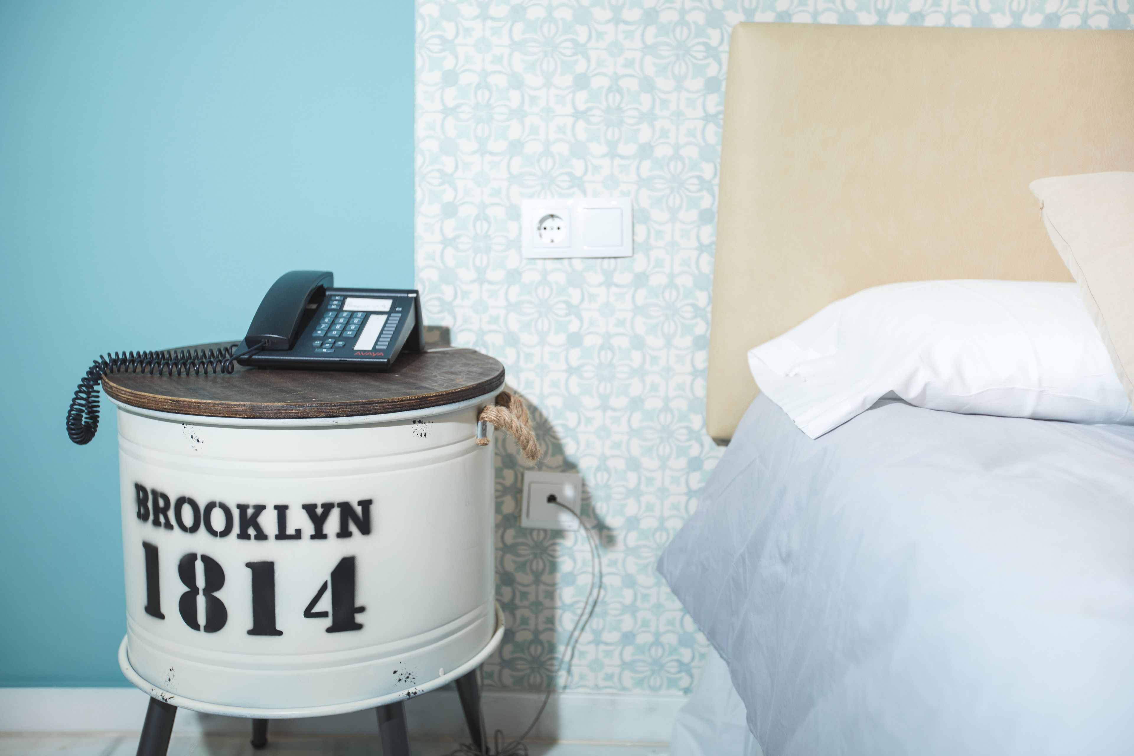 Una stanza con un letto, un oggetto cilindrico bianco con la scritta 'BROOKLYN 1814' e un telefono sopra.
