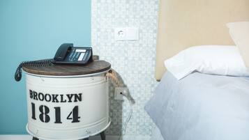 Una stanza con un letto, un oggetto cilindrico bianco con la scritta 'BROOKLYN 1814' e un telefono sopra.