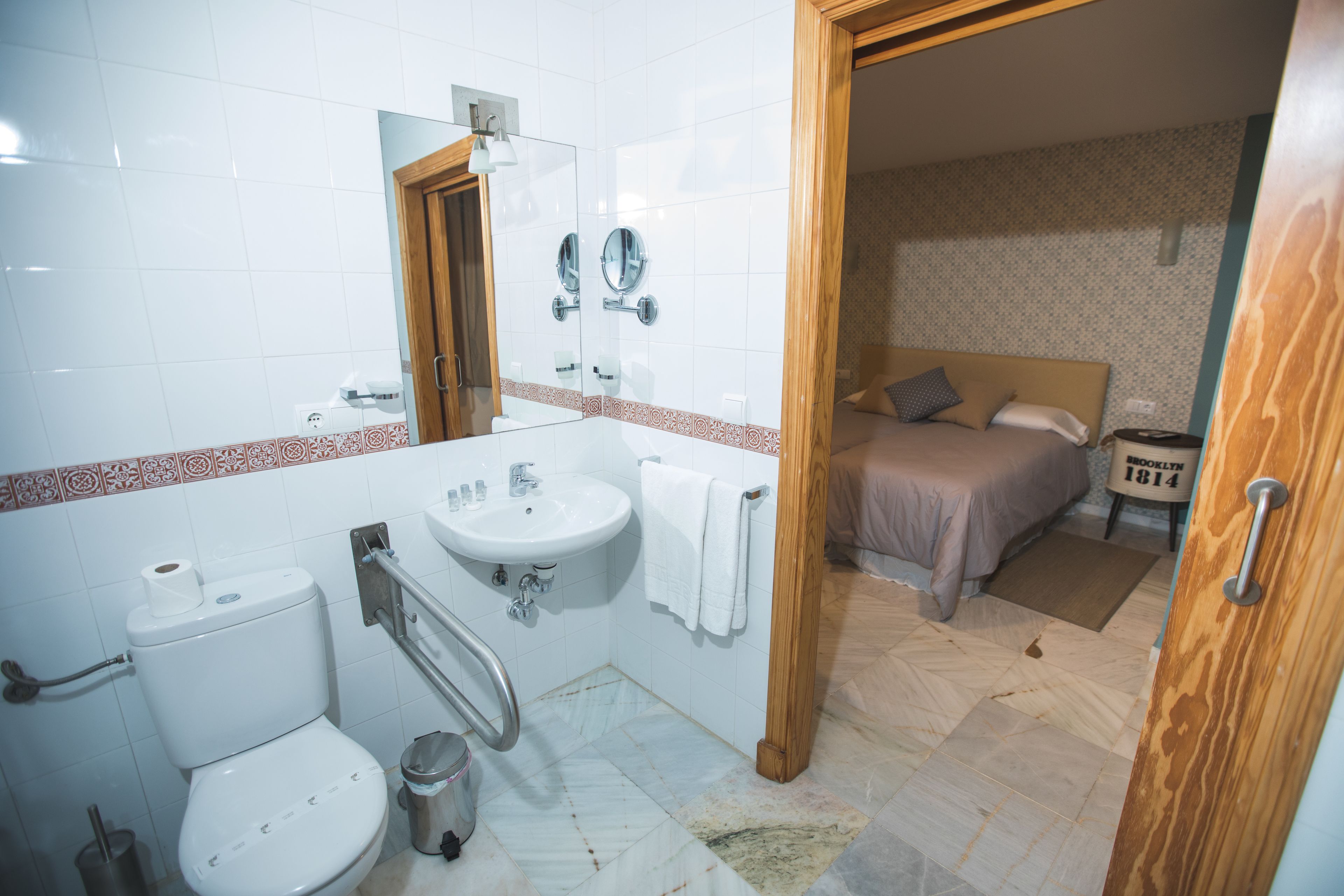 A bathroom with a toilet, sink, and mirror. There is a wooden door leading to a bedroom with a bed and a nightstand.
