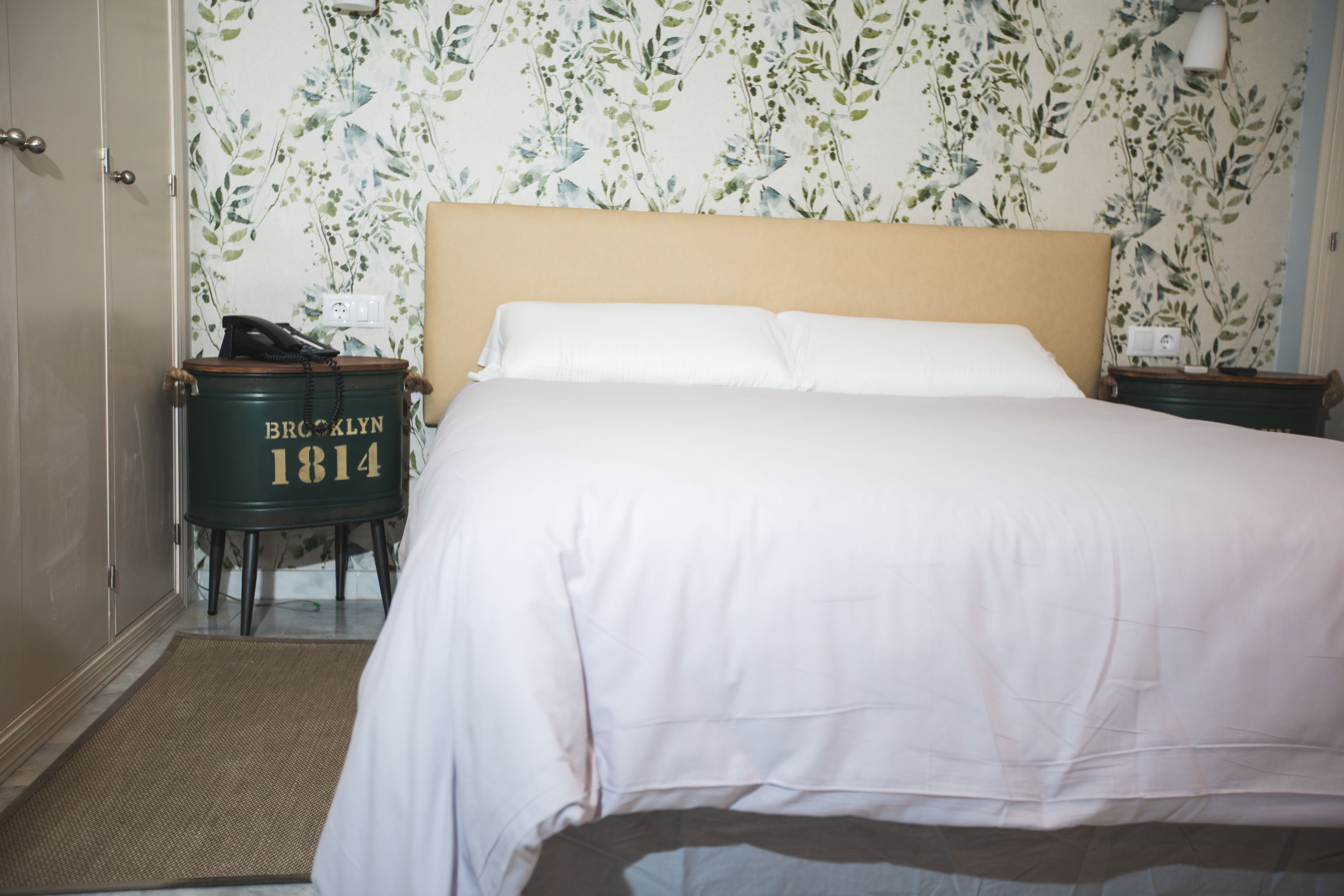 Una cama bien hecha con sábanas blancas, cabecera de madera y un estampado floral en el papel tapiz.