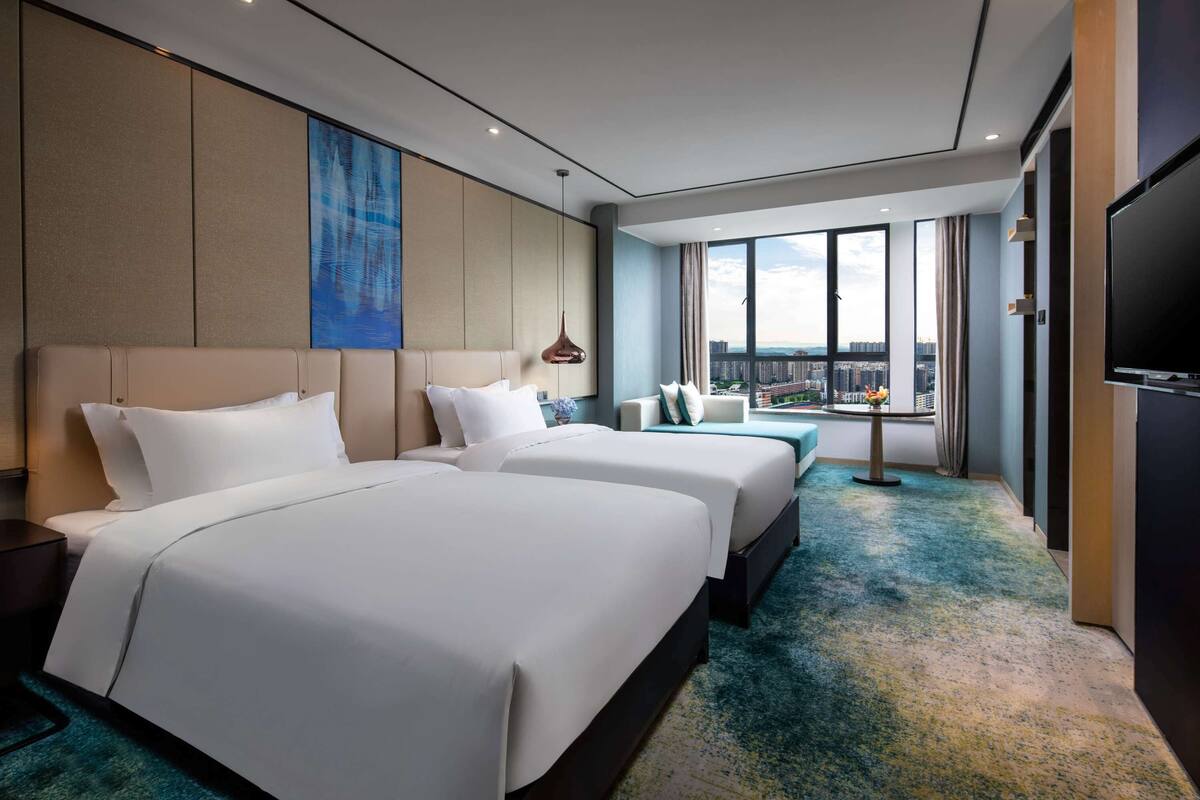 A hotel room with two beds, a TV, a large window with a city view, and a blue abstract painting on the wall.