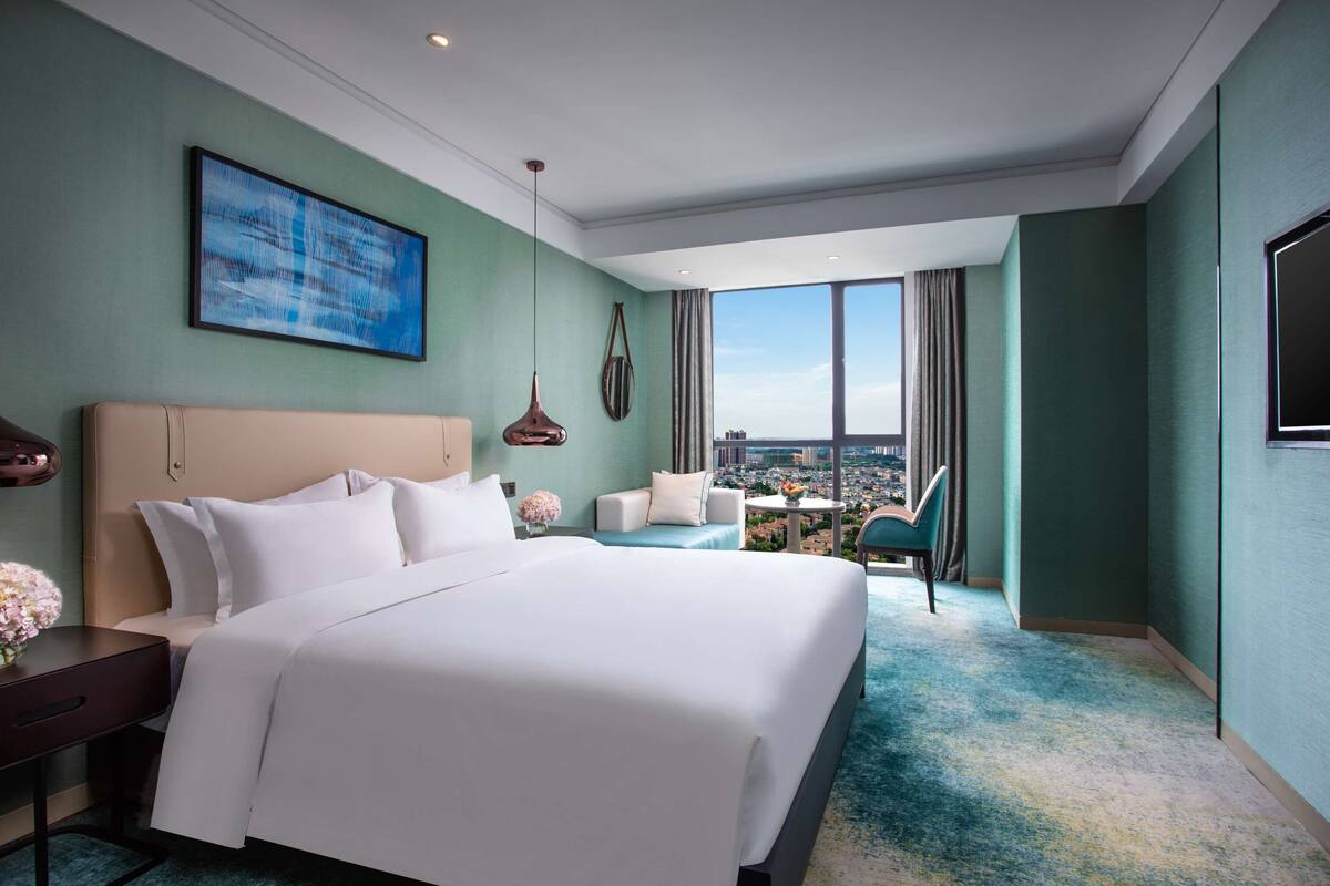 A hotel room with a large bed, a seating area with a view, and a cityscape visible through the window.