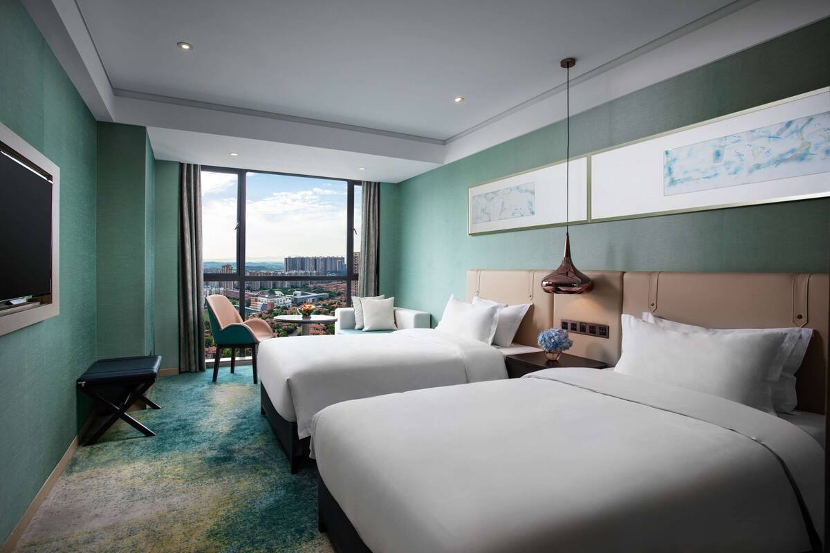 A hotel room with two beds, a desk, a chair, a TV, and a large window offering a city view.