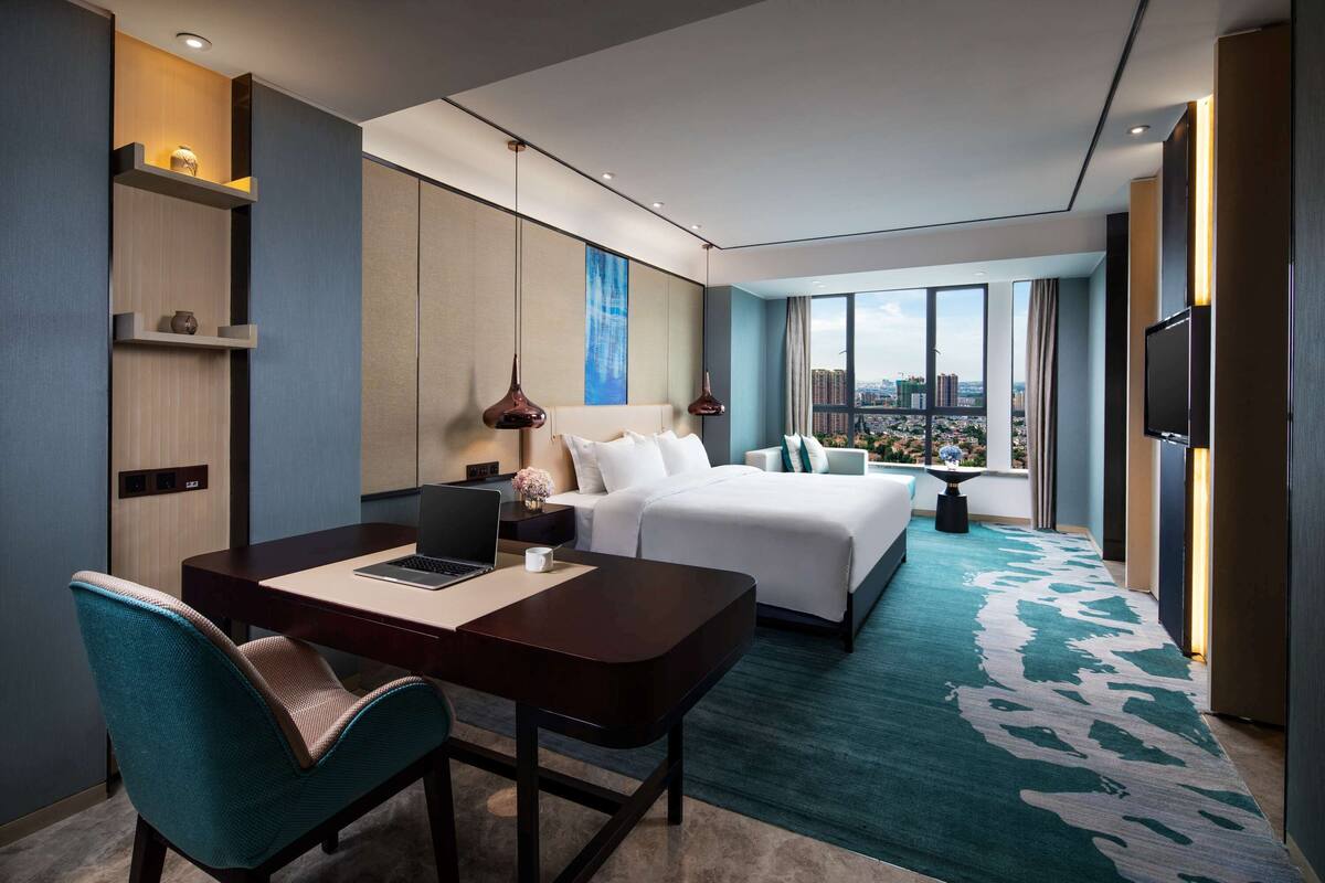 A modern hotel room with a large bed, a desk with a laptop, a chair, and a city view through large windows.