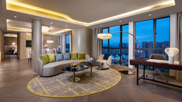 Una sala de estar moderna con un sofá curvo, una mesa de centro redonda y un escritorio con una silla. Amplias ventanas ofrecen vista a la ciudad.