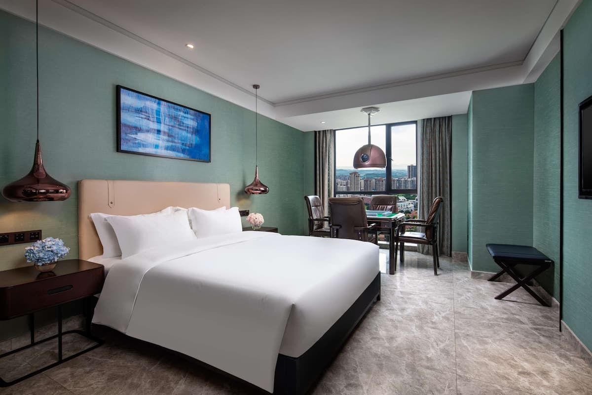 A hotel room with a large bed, a desk with a chair, a view of the city through a large window, and a painting on the wall.