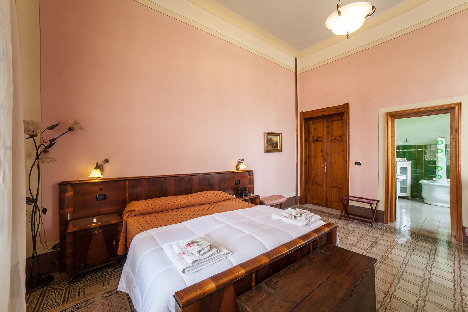 Quarto com cama de madeira, mesa de cabeceira, luminária e vista para o banheiro por uma porta aberta.